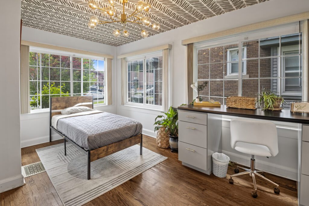 Beautiful sunroom in a historic home, full of windows, a twin bed and an office desk with natural wood floors, grey walls and white trim. Wallpapered ceiling and edison light chanedlier.